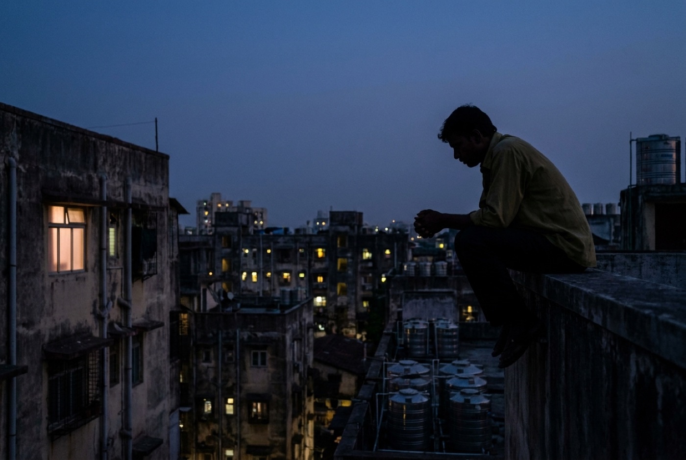 A man sitting alone on a rooftop at dusk, seen from behind, city lights in the distance